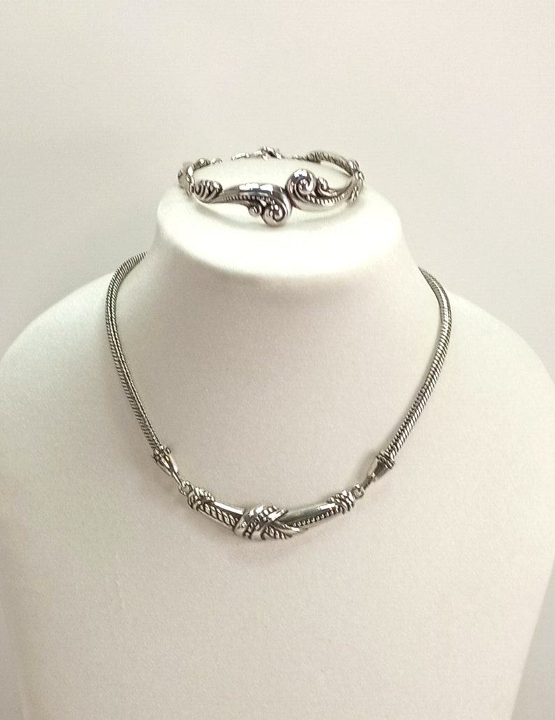 Op de afbeelding: Een zilveren ketting- en armbandset met een gedraaid touwontwerp en sierlijke details. De ketting heeft een gebogen vorm en de armband heeft een swirl-ontwerp.