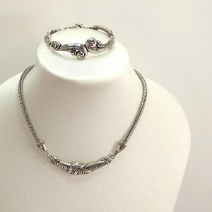 Op de afbeelding: Een zilveren ketting- en armbandset met ingewikkelde, draaiende ontwerpen. De ketting is een delicate ketting met een gebogen hanger, terwijl de armband een enkele, open band is.