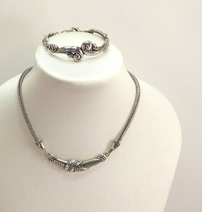 Op de afbeelding: Een zilveren ketting- en armbandset met ingewikkelde, draaiende ontwerpen. De ketting is een delicate ketting met een gebogen hanger, terwijl de armband een enkele, open band is.