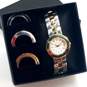 May include: A two-tone gold and silver wristwatch with a white face and black hands. The watch is in a black box with three silver rings.