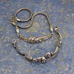 Op de afbeelding: Twee zilveren kettingen, een met een touwketting en een decoratieve zilveren hanger, de andere met een meer versierde zilveren hanger.