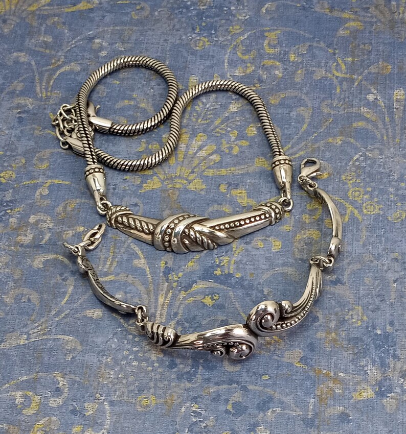 Op de afbeelding: Twee zilveren kettingen, een met een touwketting en een decoratieve zilveren hanger, de andere met een meer versierde zilveren hanger.