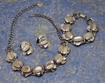 Puede incluir: Un conjunto de collar, pulsera y pendientes de tono plateado. El collar y la pulsera tienen una cadena con eslabones ovalados que tienen un diseño texturizado. Los pendientes son simples formas ovaladas con el mismo diseño texturizado.
