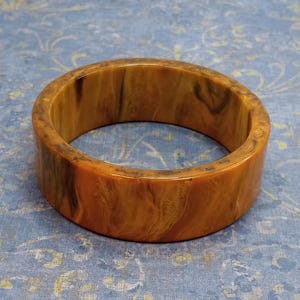 May include: A wide, vintage bangle bracelet in shades of amber, brown, and tan. The thick, circular bracelet has a marbled appearance with a glossy finish. The jewellery piece is likely made of a resin or plastic material.