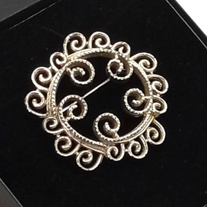 Puede incluir: Un broche de filigrana de plata con un diseño circular que presenta intrincados remolinos y un pasador en la parte posterior.