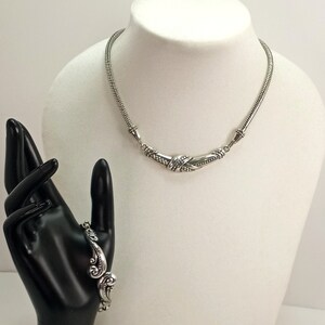 Op de afbeelding: Een zilveren ketting- en armbandset met ingewikkelde ontwerpen. De ketting heeft een gebogen, gedraaid ontwerp en de armband heeft een bloemmotief.