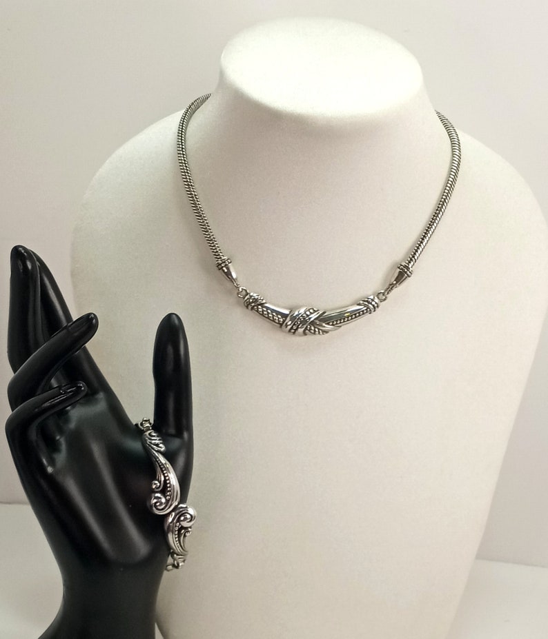 Op de afbeelding: Een zilveren ketting- en armbandset met ingewikkelde ontwerpen. De ketting heeft een gebogen, gedraaid ontwerp en de armband heeft een bloemmotief.