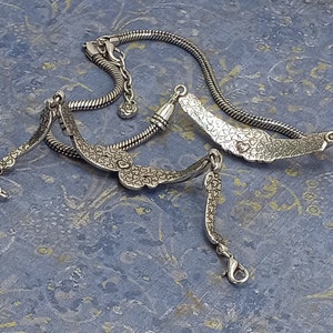 Op de afbeelding: Een zilveren kettingarmband met een hartvormige bedel. De bedel is versierd met kleine hartjes en heeft een hangend hartje.