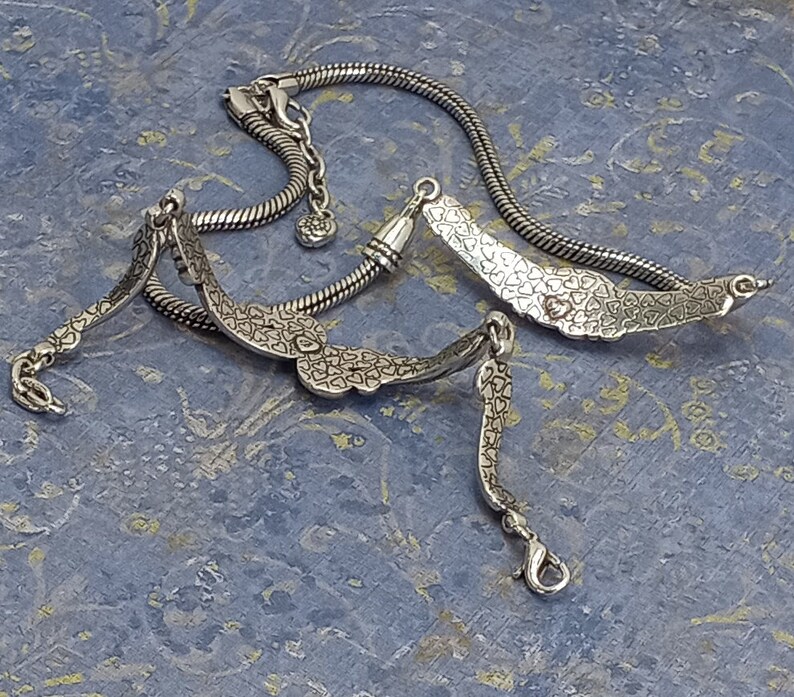 Op de afbeelding: Een zilveren kettingarmband met een hartvormige bedel. De bedel is versierd met kleine hartjes en heeft een hangend hartje.