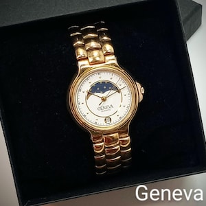 May include: Gold-toned Geneva Quartz watch with a white face featuring a moon phase design and a date display. The watch has a gold band and is presented in a black box. The word "Geneva" is printed on the watch face.