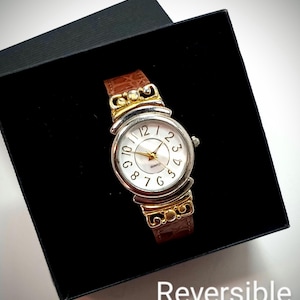 Puede incluir: Un reloj de cuarzo plateado y dorado con esfera blanca y correa de cuero marrón. El reloj se muestra en una caja negra. La palabra "Reversible" está impresa en la parte inferior de la imagen.