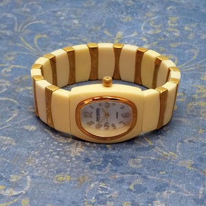 May include: Cream quartz watch with gold-tone bezel and band accents. The watch face has a mother-of-pearl effect with gold hands and numerals. The band is made of cream segments with gold connectors. A stylish timepiece.