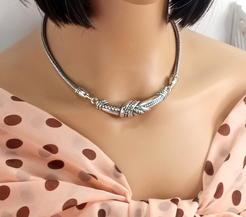 Op de afbeelding: Een zilveren choker ketting met een gedraaid, getextureerd ontwerp. De ketting is gemaakt van een dunne ketting en heeft een decoratieve sluiting.