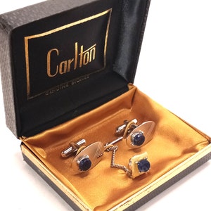 Könnte beinhalten: Ein Set Manschettenknöpfe und ein Krawattennadel in einer schwarzen Box mit goldenen Verzierungen. Die Manschettenknöpfe und die Krawattennadel haben blaue Steine und goldene Akzente. Die Box ist mit goldenem Satin ausgekleidet und trägt innen den Schriftzug "Carlton Genuine Stones".