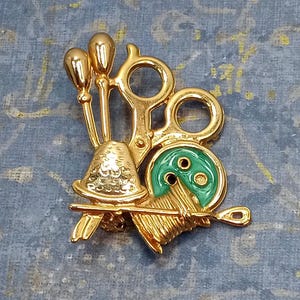May include: Gold-toned brooch featuring a sewing theme with a pair of scissors, a spool of thread, a thimble, and a button with green enamel.