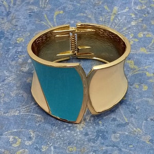 May include: A gold bangle bracelet with a turquoise and cream enamel design. The bracelet is open and has a hinge closure.