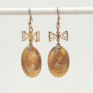 以下が含まれることがあります： 金色のイヤリングのペア。各イヤリングは、透明なクリスタルが付いた弓型のアクセント、小さな丸いクリスタル、テクスチャードデザインの楕円形のペンダントが特徴です。イヤリングは金色のフックから吊り下げられています。