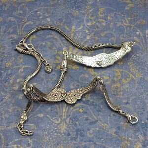 Op de afbeelding: Twee zilveren armbanden met een hartvormig ontwerp. De armbanden zijn aan elkaar gekoppeld en hebben een kettingachtig ontwerp.