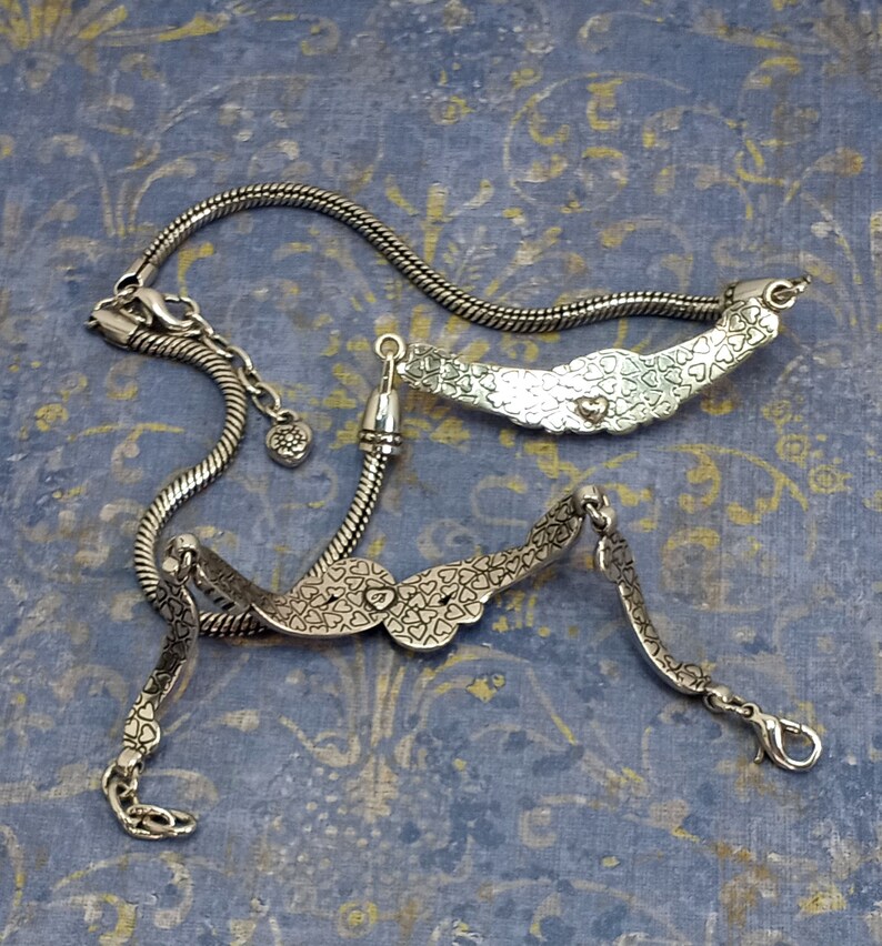 Op de afbeelding: Twee zilveren armbanden met een hartvormig ontwerp. De armbanden zijn aan elkaar gekoppeld en hebben een kettingachtig ontwerp.