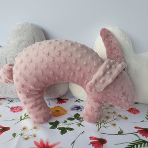 Puede incluir: Una almohada de viaje de felpa rosa y blanca con forma de conejo. La almohada est&aacute; sobre un fondo blanco con un patr&oacute;n floral.