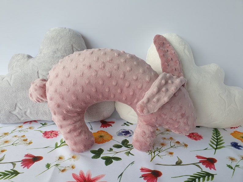 Puede incluir: Una almohada de viaje de felpa rosa y blanca con forma de conejo. La almohada est&aacute; sobre un fondo blanco con un patr&oacute;n floral.