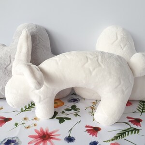 Puede incluir: Una almohada de peluche blanca con forma de conejo con un patr&oacute;n de estrellas grises. La almohada est&aacute; sobre un fondo blanco con un patr&oacute;n floral.