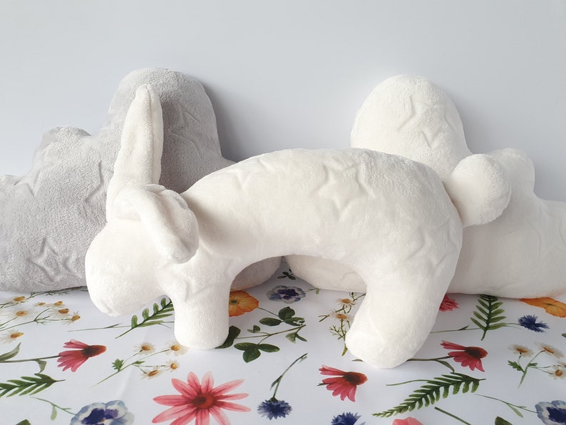 Puede incluir: Una almohada de peluche blanca con forma de conejo con un patr&oacute;n de estrellas grises. La almohada est&aacute; sobre un fondo blanco con un patr&oacute;n floral.
