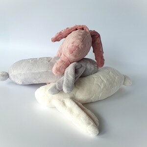 Puede incluir: Tres juguetes de peluche en forma de conejo de diferentes colores: rosa, gris y blanco. El conejo rosa est&aacute; encima del conejo gris, y el conejo blanco est&aacute; en la parte inferior.