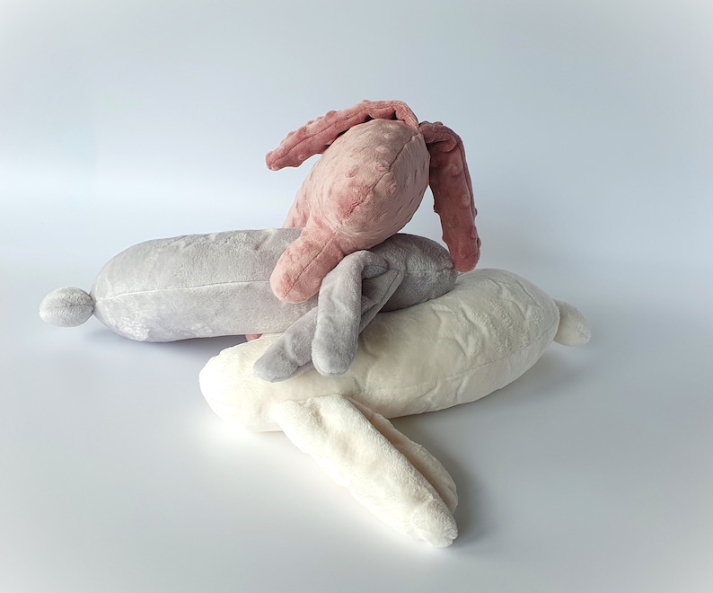 Puede incluir: Tres juguetes de peluche en forma de conejo de diferentes colores: rosa, gris y blanco. El conejo rosa est&aacute; encima del conejo gris, y el conejo blanco est&aacute; en la parte inferior.