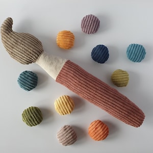 Könnte beinhalten: Ein Plüsch-Pinselspielzeug aus braunem und terrakottafarbenem Kordstoff mit einem cremefarbenen Mittelband, umgeben von neun kleinen, runden, bunten Kordbällen in den Farben Violett, Orange, Blau, Türkis, Olivgrün, Gelb und Rosa.