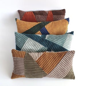 Op de afbeelding: Een stapel van vijf rechthoekige corduroy kussens in verschillende kleuren. De kussens hebben geometrische patchwork ontwerpen in tinten van oranje, bruin, groen, blauw en geel. De kussens zijn verticaal geplaatst tegen een witte achtergrond.