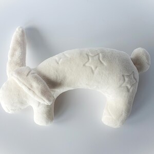 Puede incluir: Una almohada de cuello de peluche blanca con forma de conejo con bordados en forma de estrella.