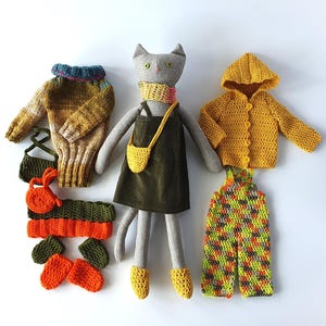 Op de afbeelding: Een grijze stoffen kattenpop met een donkergroene corduroy jurk, een gele gehaakte sjaal, een gele schoudertas en gele slofjes. Accessoires zijn onder meer een gebreide trui, een geel vest met capuchon, een tuinbroek en kleine gehaakte items.