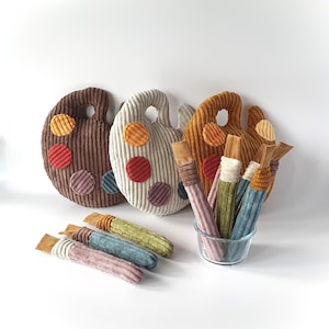 Puede incluir: Tres cojines de pana con forma de paletas de artista en marrón oscuro, crema y amarillo mostaza, cada uno con parches de tela circulares de varios colores. Varios pinceles de peluche de pana de colores surtidos están dispuestos cerca.