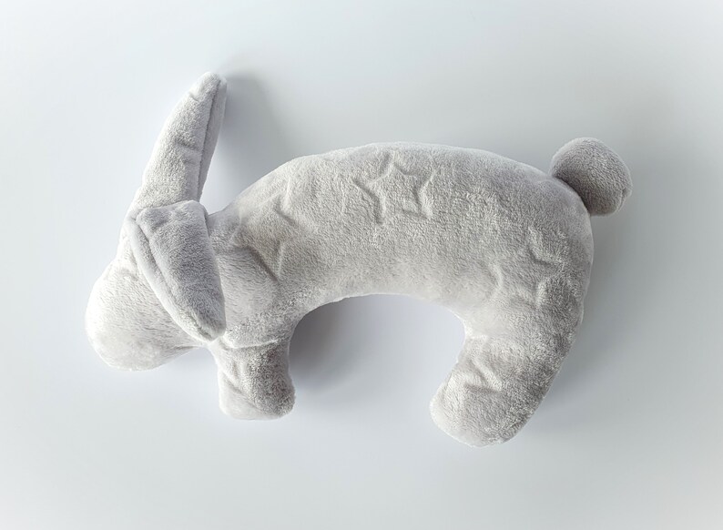 Puede incluir: Una almohada de cuello con forma de conejo de peluche gris con un patr&oacute;n de estrellas.