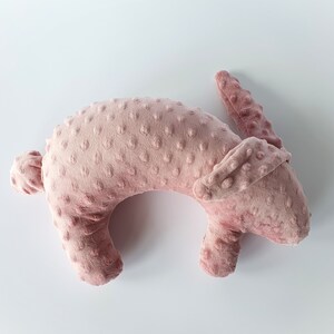 Puede incluir: Una almohada de peluche rosa en forma de conejo con una superficie texturizada. La almohada est&aacute; tumbada de lado con la cabeza girada hacia la derecha.