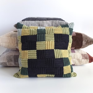 Könnte beinhalten: Ein Stapel Cord-Zierkissen in verschiedenen Farben. Das vordere Kissen hat ein gewebtes Muster in Schwarz, Grün und Gelb. Weitere Kissen sind in Grau-, Beige- und Brauntönen gehalten. Die Kissen sind quadratisch und wirken weich und bequem.