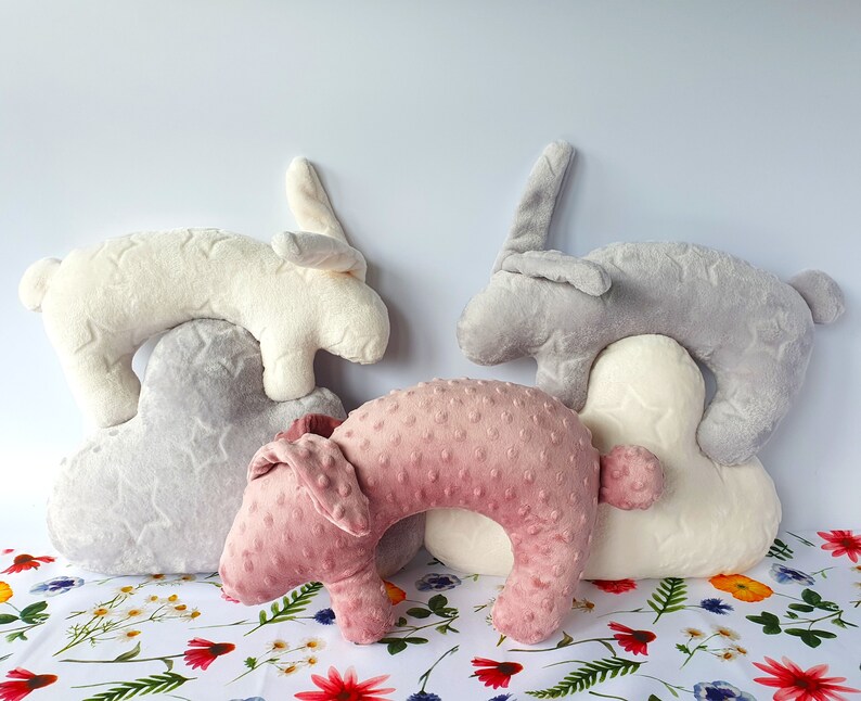 Puede incluir: Tres almohadas de peluche con forma de conejo en gris, blanco y rosa. Las almohadas est&aacute;n apiladas, con el conejo gris en la parte superior, el conejo blanco en el medio y el conejo rosa en la parte inferior. Las almohadas est&aacute;n hechas de un tejido suave y peludo y tienen un patr&oacute;n de estrellas.