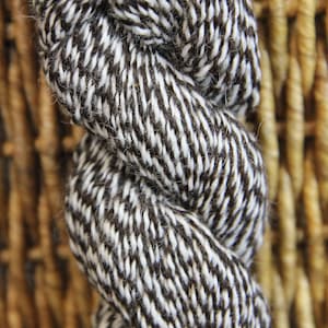 May include: Close-up of a twisted skein of yarn. The yarn is a mix of black and white fibers, creating a textured, diagonal pattern. The background is a woven, light brown material.