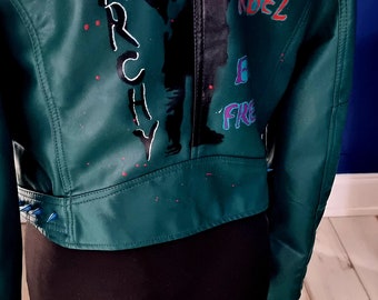 Könnte beinhalten: Eine grüne Lederjacke mit einer schwarzen Silhouette einer Person mit Flügeln und den Worten "Stay Rude", "Rebel", "Free" und "Anarchy", die darauf gemalt sind. Die Jacke hat blaue Spikes am Kragen.