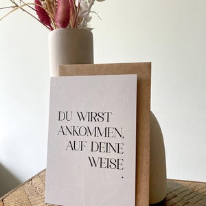 Könnte beinhalten: Eine Grußkarte mit dem Text "DU WIRST ANKOMMEN, AUF DEINE WEISE" in schwarzer Schrift, mit floralem Design. Die Karte wird mit einem braunen Umschlag kombiniert und vor einer beigen Vase mit Trockenblumen platziert.