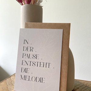Könnte beinhalten: Eine Grußkarte mit dem Text "IN . DER . PAUSE . ENTSTEHT . DIE . MELODIE" in schwarzer Schrift, mit floralem Design. Die Karte liegt vor einem braunen Umschlag und einer beigen Vase mit Trockenblumen.