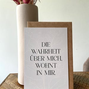 Può includere: Un biglietto di auguri con il testo "DIE WAHRHEIT ÜBER MICH, WOHNT IN MIR." in nero su sfondo bianco con un motivo floreale. Il biglietto è posto davanti a una busta marrone e un vaso beige con fiori secchi.