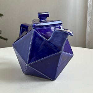 Può includere: Una teiera geometrica e sfaccettata in una tonalità blu intenso. La teiera in ceramica presenta un coperchio quadrato con una piccola maniglia e un beccuccio. Il design è moderno e angolare, con una finitura lucida. La teiera è un pezzo suggestivo per servire il tè.