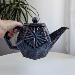 Può includere: Teiera in ceramica blu scuro con motivo floreale geometrico. La teiera ha un coperchio con un piccolo pomello e un lungo beccuccio. Il manico è angolare. La teiera è tenuta da una mano.