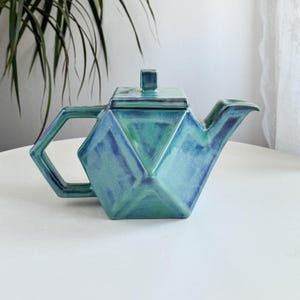 Può includere: Teiera geometrica in ceramica nei toni del turchese e del blu. La teiera presenta un design sfaccettato con coperchio e manico quadrati. Il beccuccio è curvo. La teiera è su una superficie bianca.