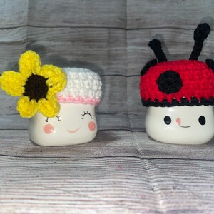 May include: Two ceramic mugs with smiling faces. One mug has a white and pink crocheted rim with a yellow and brown sunflower. The other mug has a red and black crocheted ladybug hat with black antennae. The mugs are on a light wood-grain surface.