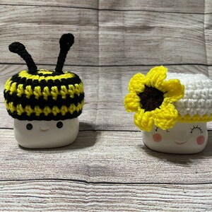 May include: Two white ceramic mugs with smiling faces, each adorned with a crocheted hat. One mug features a black and yellow striped bee hat with antennae, and the other has a white hat with a yellow sunflower.