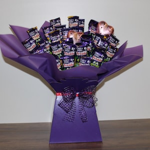 May include: A purple gift box filled with a bouquet of chocolate bars wrapped in purple foil. The chocolate bars are Dairy Milk and Freddo. There are two pink heart-shaped decorations on top of the bouquet.