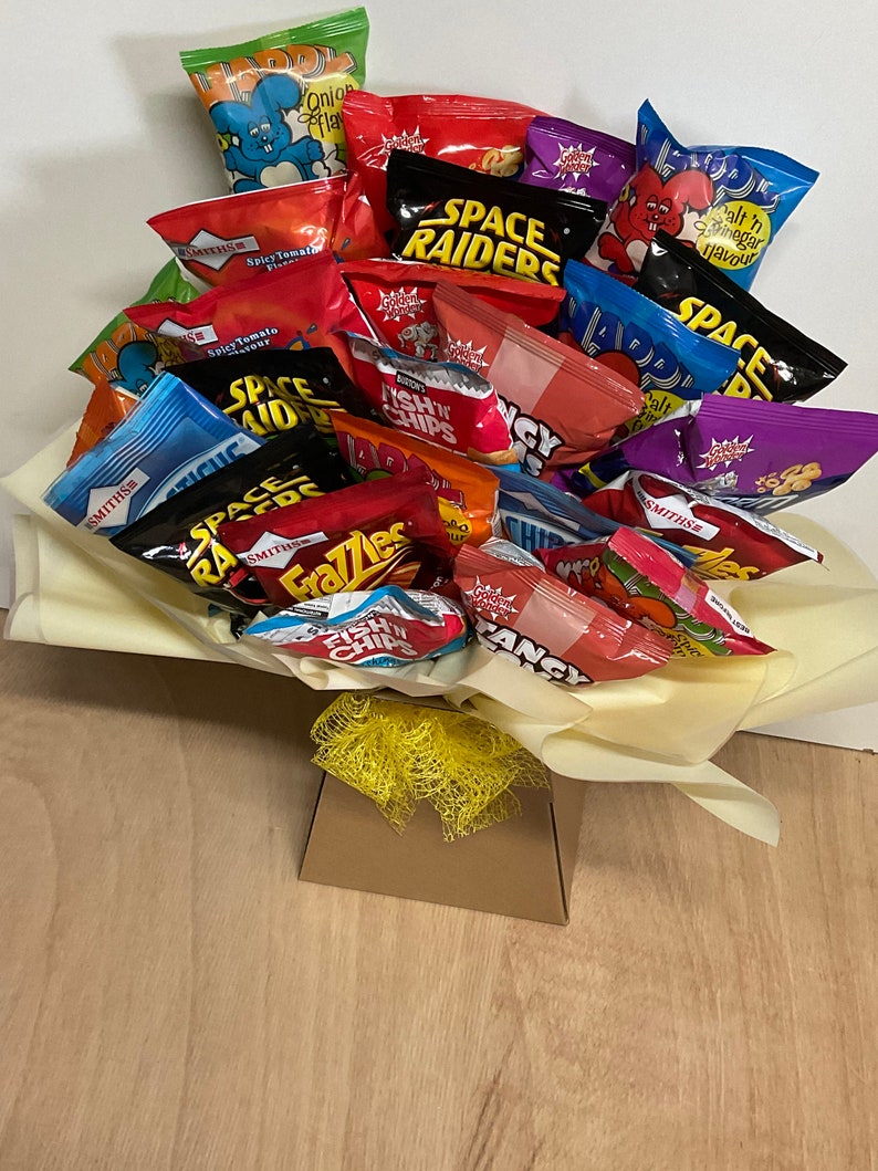 May include: A bouquet of various snack chip bags, including brands like Space Raiders, Frazzles, and Fish 'n' Chips. The bags are arranged in a decorative display, with a yellow bow and a brown box at the base. The chips are in colorful packaging.